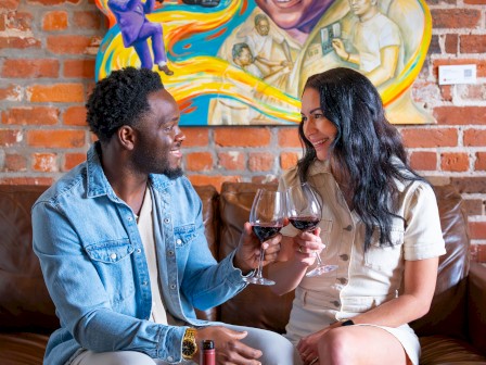 Two people share a toast on a couch, wine glasses raised, a bottle on the table, with a bright vinyl mural of jazz legends on a brick wall.