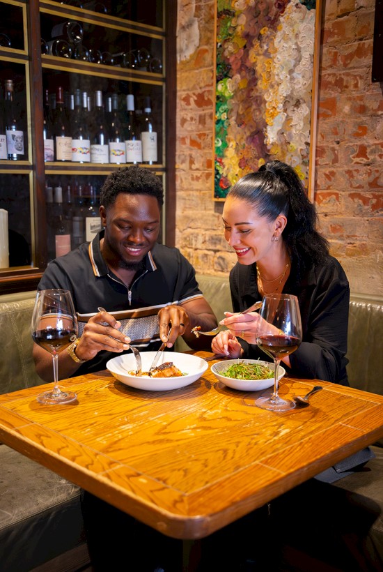 Two people are dining at a restaurant with wine, sharing a meal, seated at a wooden table with wine bottles in the background.
