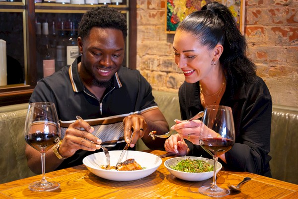 Two people enjoying a meal with wine at a restaurant. The table has dishes and glasses, and the background shows shelves with bottles.