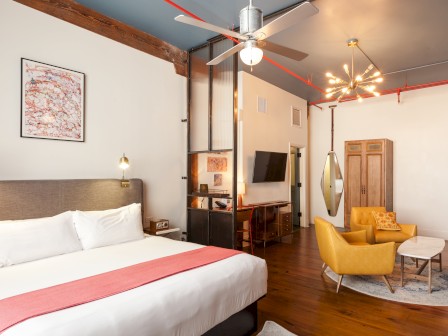 A stylish hotel room featuring a bed, seating area with yellow chairs, TV, ceiling fan, and modern lighting.