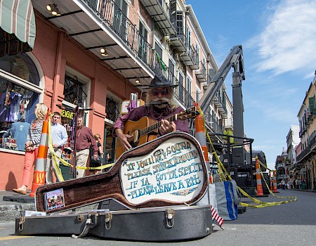 A street performer playing guitar next to an open guitar case with a humorous sign in a lively urban setting.