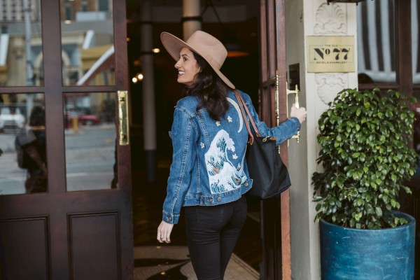 A person in a hat and denim jacket enters a building with a large plant nearby and a chandelier inside, creating a stylish scene.