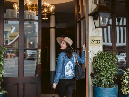 A person wearing a hat and denim jacket is entering a building with large glass doors, surrounded by potted plants.