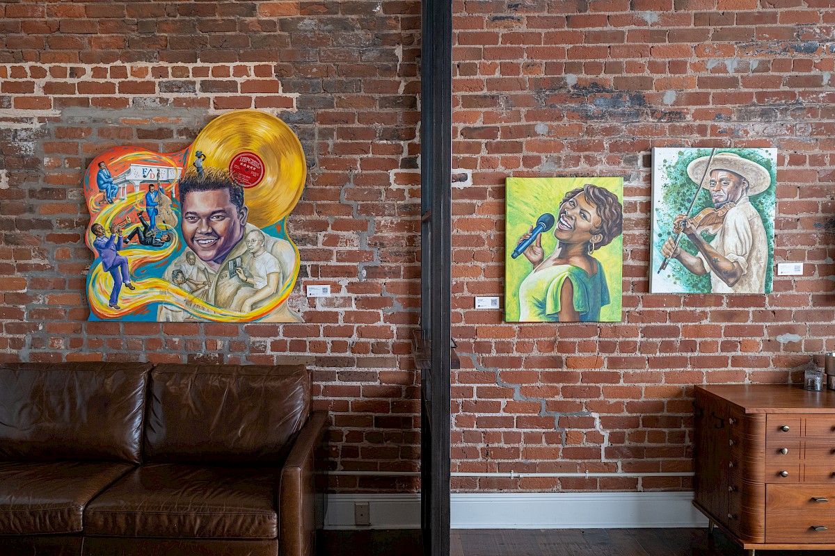 The image shows artwork on a brick wall featuring portraits, a leather couch, and a wooden cabinet in a cozy room setting.