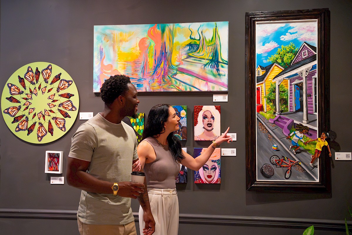 A couple observes vibrant artwork in a gallery, featuring colorful abstract and figurative paintings on the wall.