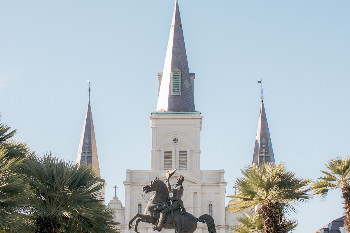 A statue of a rider on a horse with a cathedral and palm trees in the background under a clear blue sky.