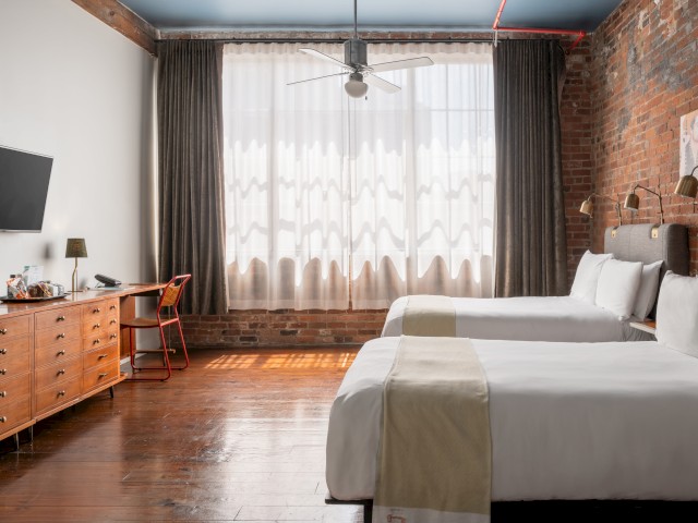 The image shows a hotel room with two beds, a TV, a desk with a chair, large window curtains, and a rustic brick wall.