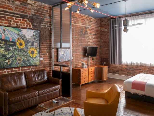 The image shows a stylish room with brick walls, a leather sofa, a bed, modern lighting, and a sunflower painting on the wall.