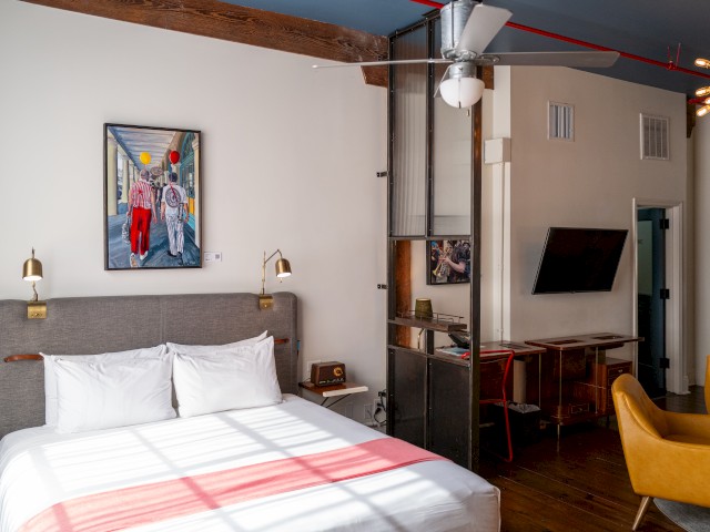 A modern bedroom with a bed, artwork, ceiling fan, TV, two chairs, and desk. Wood beams and a red-striped bed cover add warmth.