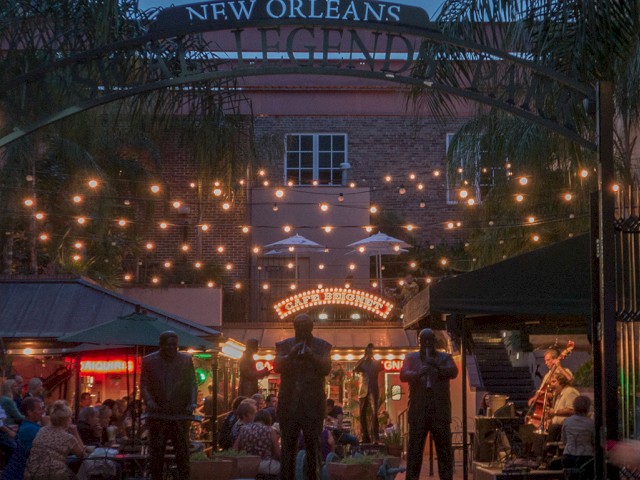 A lively New Orleans courtyard scene, with string lights, people dining, and a musician performing under a sign saying 