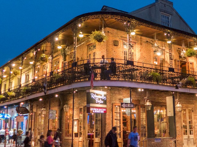 People walking past a lively, illuminated brick building with a balcony, plants, and patio seating at dusk.