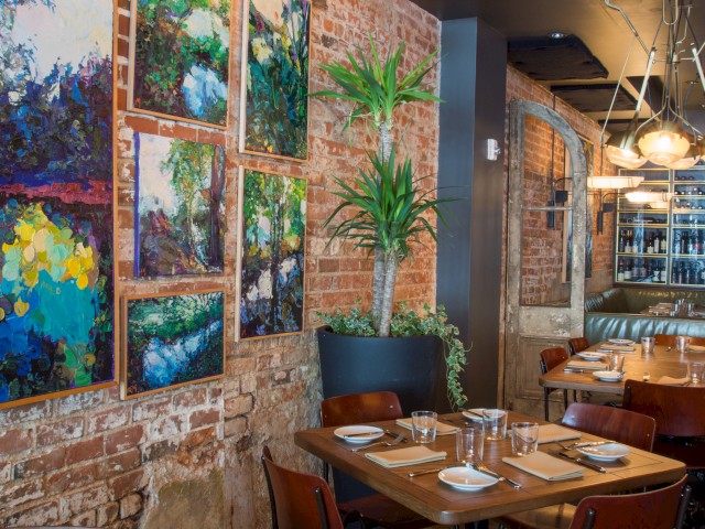 A cozy restaurant interior with brick walls, colorful paintings, modern lighting, and tables set for dining.