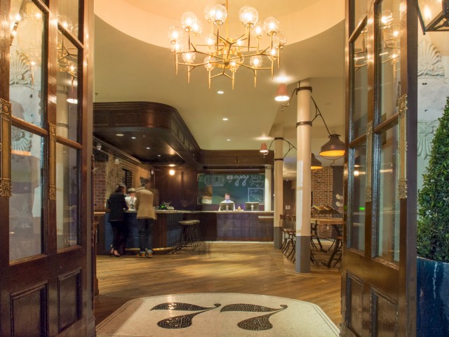 An elegant hotel lobby with open doors, chandelier lighting, reception desk, and people standing by it, featuring a warm, inviting atmosphere.