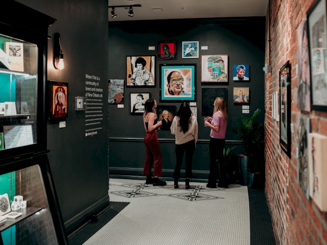 Three people observe art on a black wall in a gallery, with portraits and photos displayed.
