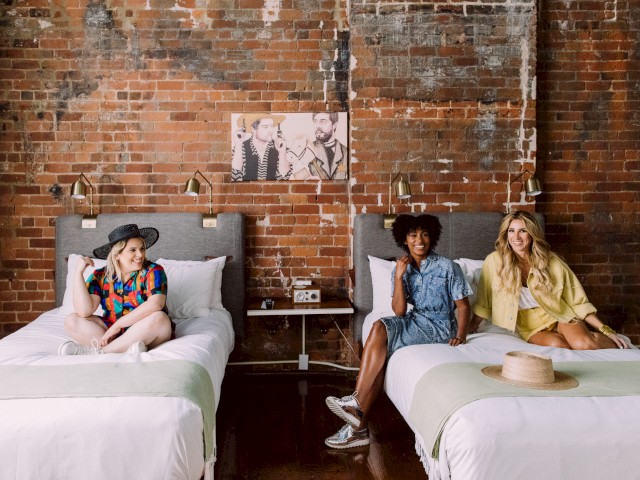 Three people sitting on beds in a room with brick walls and artwork, enjoying a relaxed atmosphere.