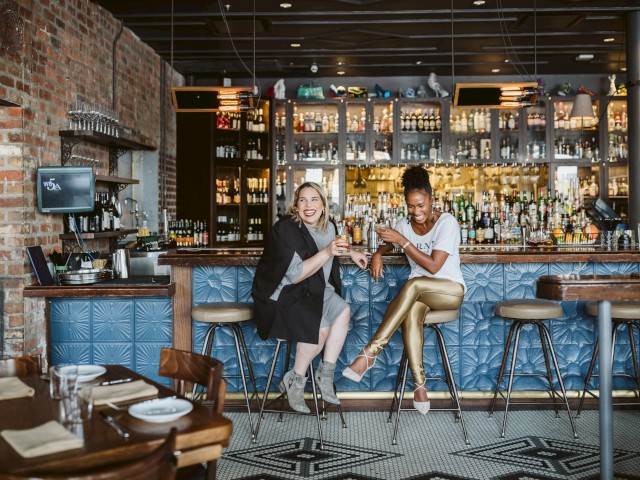 Two people sitting at a bar with drinks, surrounded by a stylish, well-stocked interior, and an empty table in the foreground.