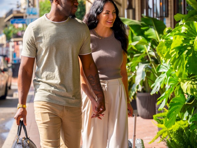 A couple walks hand in hand on a sunny street, surrounded by plants, carrying luggage.