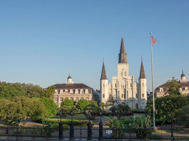 A historic building with spires, surrounded by gardens, with an American flag in the foreground under a clear blue sky.