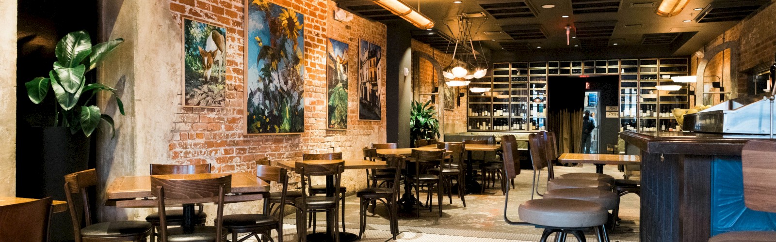 A cozy restaurant with brick walls, wooden tables, chairs, potted plants, a tiled floor, and framed artwork, creating a welcoming atmosphere.