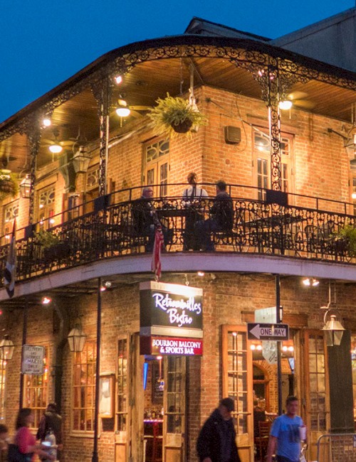 A two-story, US-style brick building with string lights and hanging plants, a bustling sidewalk, and people outside at dusk.