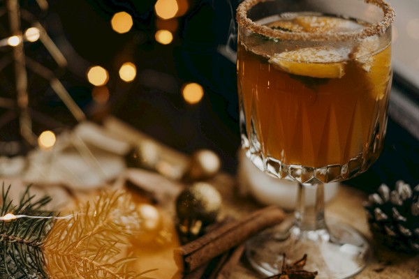 A warm drink in a glass on a wooden slice, surrounded by pine cones, cinnamon sticks, lemon slices, and festive golden lights in a cozy scene.
