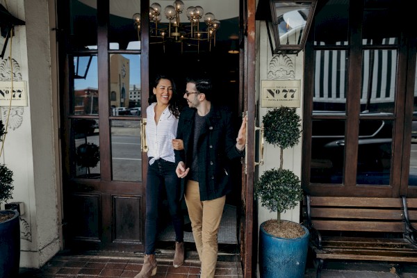 A happy couple joyfully exits a building, sharing a laugh and enjoying each other's company outside a charming entrance with plants and warm lighting.