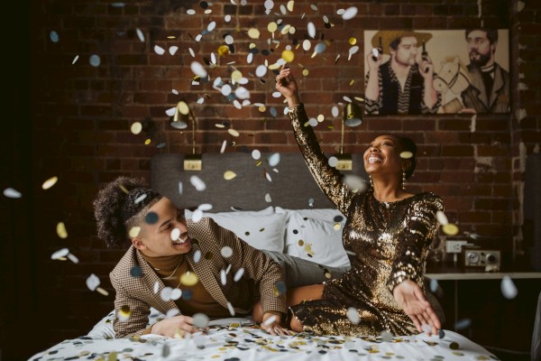 Two friends partying on a bed, tossing confetti in a cozy, stylish room; vibrant celebration vibes and playful glam.