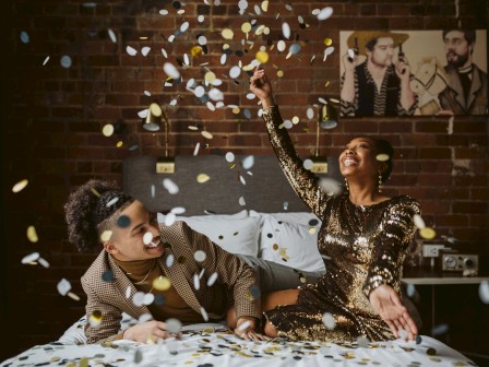Two stylish friends party on a bed as confetti rains down, one cheering with a raised champagne glow while the other leans in with a smile.