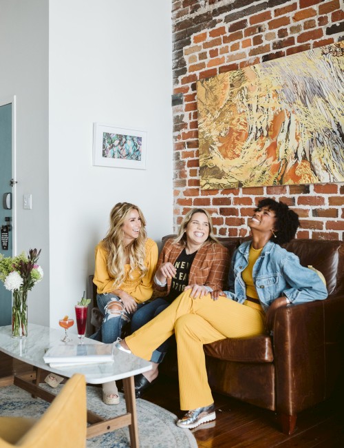 Three women laughing and enjoying each other's company on a brown leather couch in a cozy, artsy room with exposed brick wall and colorful artwork.