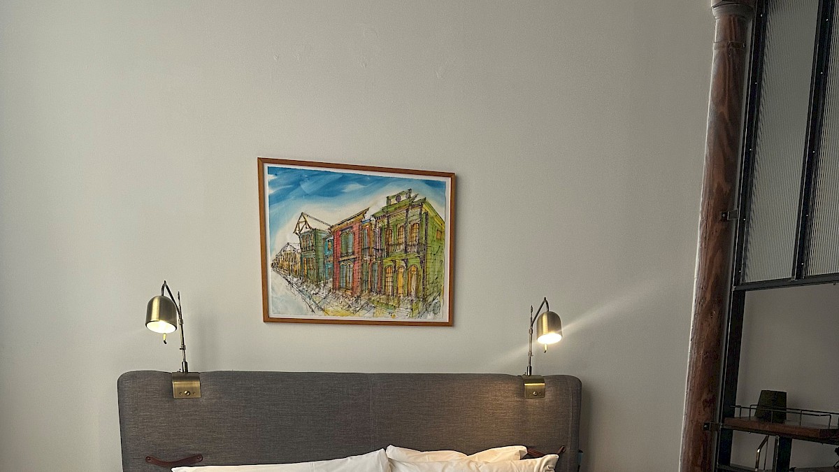 A cozy bedroom with a neatly made bed, wall-mounted lamps, colorful artwork above the headboard, and minimalistic furniture.