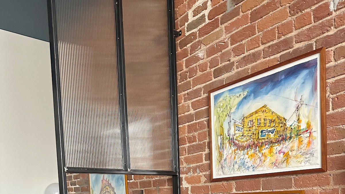 The image shows a cozy corner with exposed brick walls, framed colorful paintings, a brown leather sofa, and a wooden dresser with glass cups.