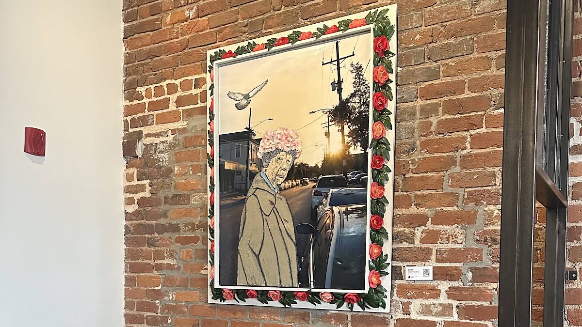 A leather sofa against an exposed brick wall features a framed artwork of an elderly woman with a floral hat, open-air sunset, parked cars, and birds.