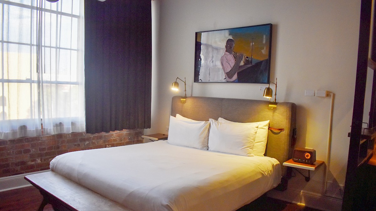 A cozy hotel room features a bed with white sheets, a wooden bench, exposed brick wall, large window, and artwork above the headboard.