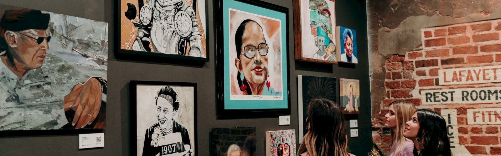 Three women are viewing and discussing artwork in an art gallery with a brick wall and a 