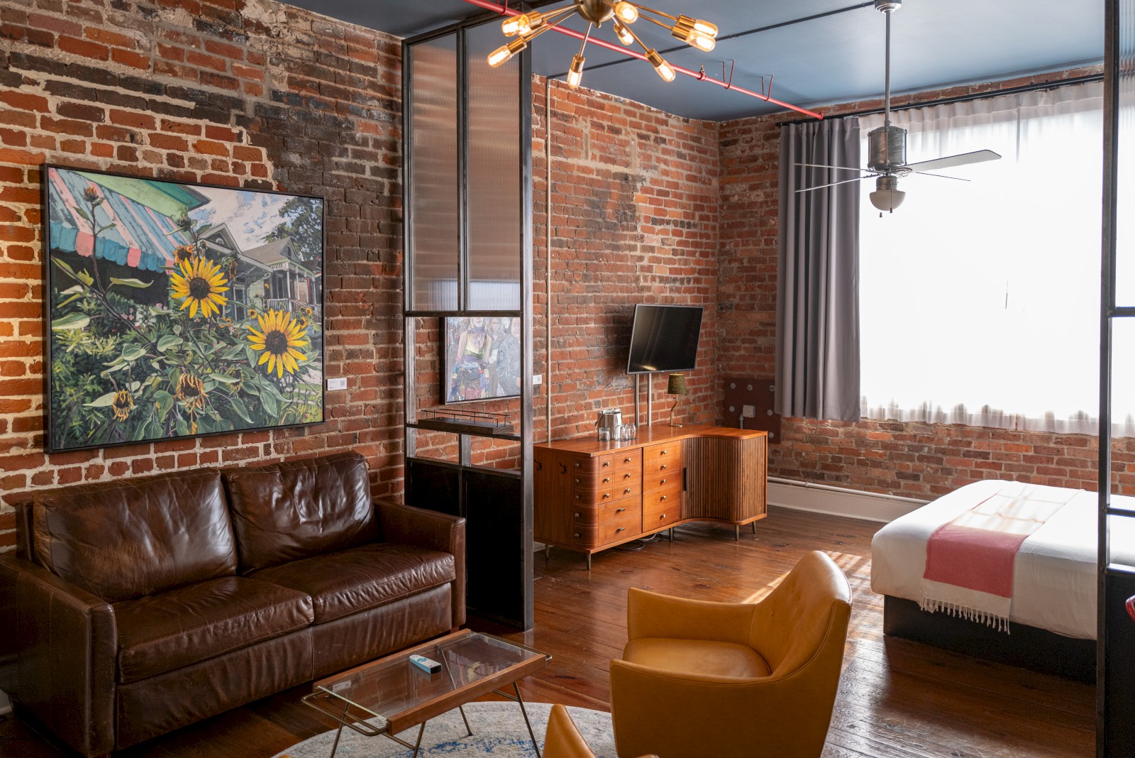 A stylish apartment with exposed brick walls, a leather sofa, modern art, a TV, and a bed near a bright window.
