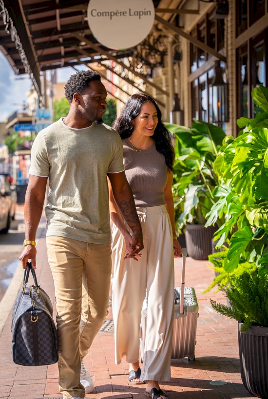A stylish couple strolls on a sunny sidewalk, holding hands, with luggage and shopping bags, surrounded by lush greenery outside a café.