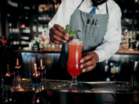 A bartender is garnishing a bright red cocktail with mint and a raspberry, in a lively bar setting with various bottles in the background.