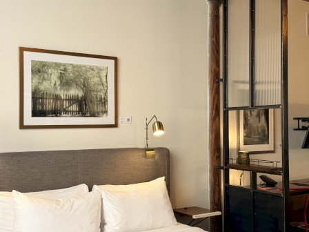 This image shows a cozy hotel room corner with a bed, framed artwork, and a metal shelving unit separating the sleeping area from the workspace.