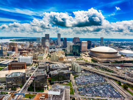 A vibrant city skyline with tall buildings, a large domed stadium, a river, and a parking lot, under a partly cloudy blue sky.