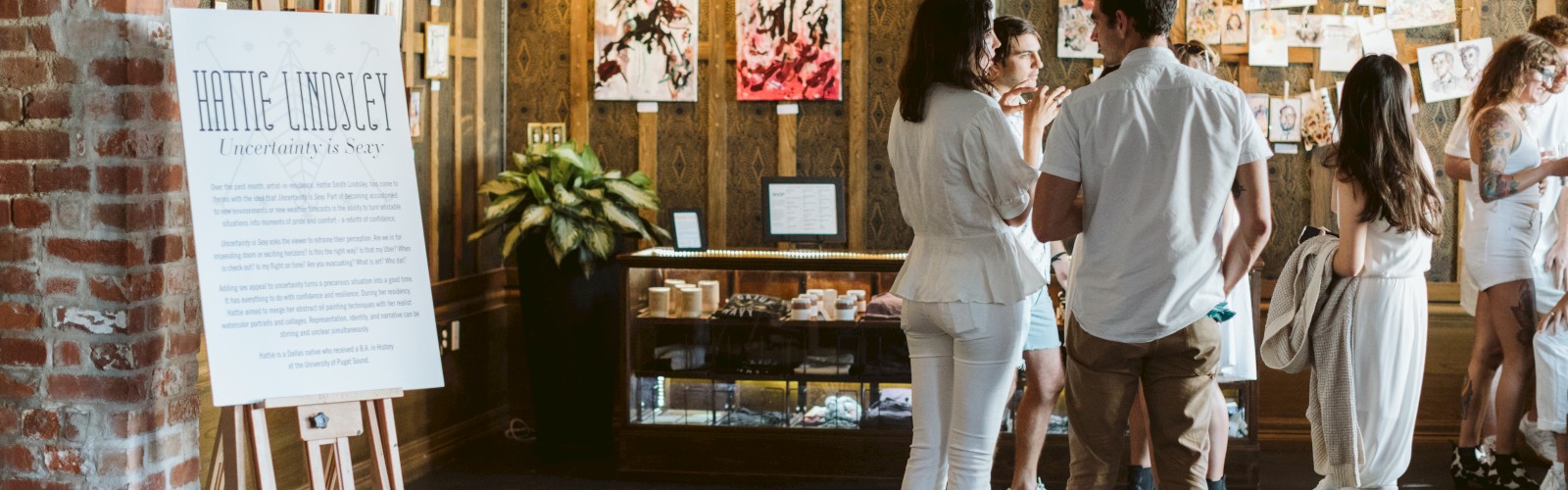 A group of people are chatting in an art gallery with paintings and drawings on the wooden walls, along with a large sign about Hattie Lunssey.