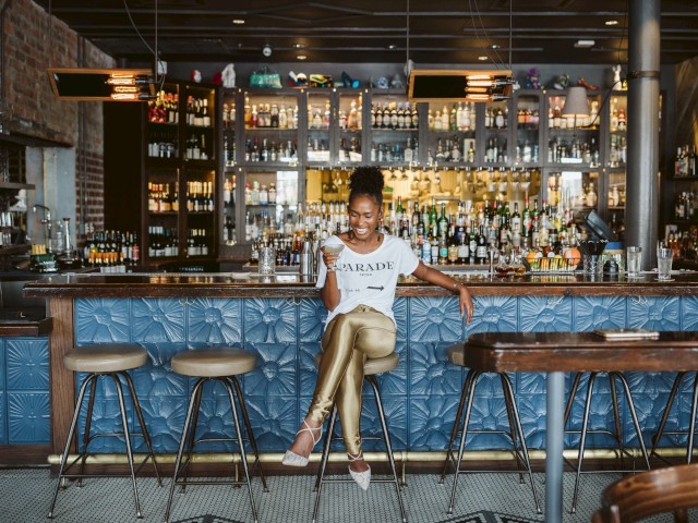 A woman is sitting at a bar, smiling and holding a cocktail, with shelves of liquor bottles behind her in a stylish restaurant.