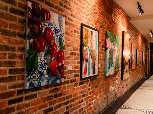 This image shows a gallery wall with various colorful paintings hanging on a red brick wall.