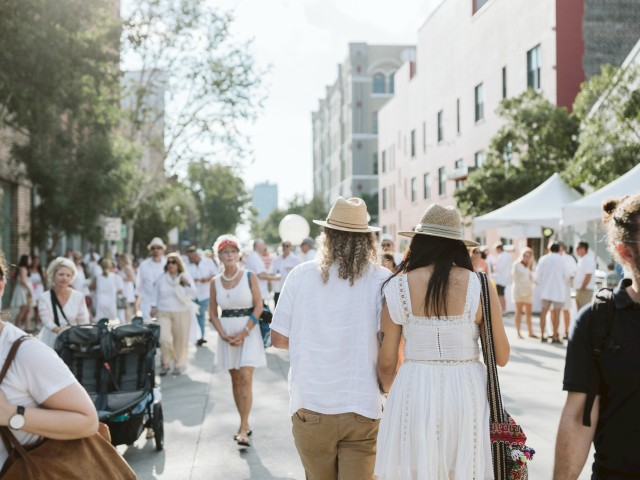 People dressed in white walk on a sunny city street with tents and buildings in the background, possibly during an outdoor event or festival.