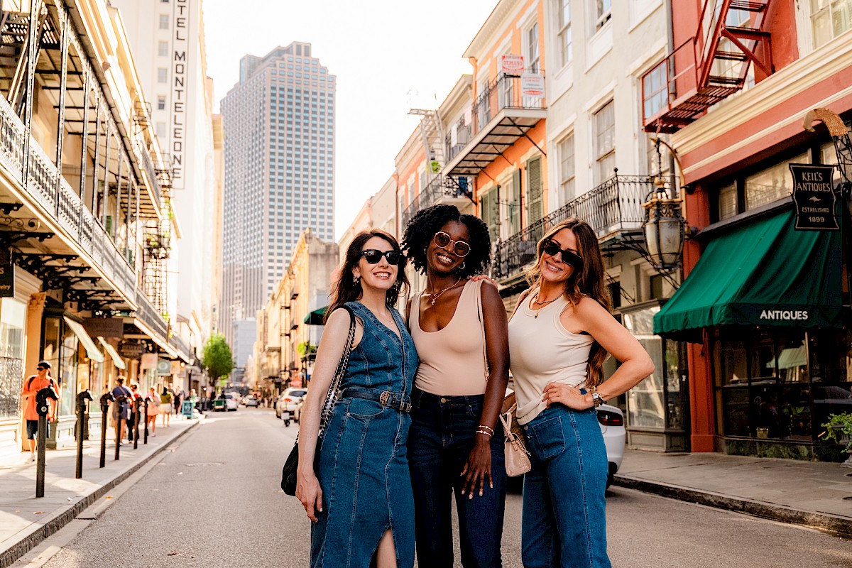 Three women pose happily on a vibrant city street, surrounded by colorful buildings and tall skyscrapers in the background.